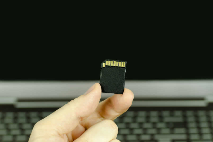 Close-up view of a hand holding a black SD memory card with a background laptop, isolated in the background