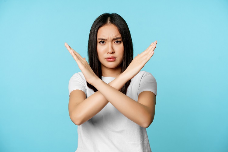 Mulher asiática com cara séria fazendo sinal de X com os braços, de pé sobre fundo azul